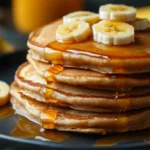 A stack of peanut butter banana pancakes topped with sliced bananas and drizzled with maple syrup, served on a black plate.