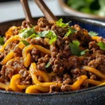 A close-up of Mongolian ground beef noodles in a dark ceramic bowl, topped with fresh cilantro and coated in a glossy sweet-savory sauce.
